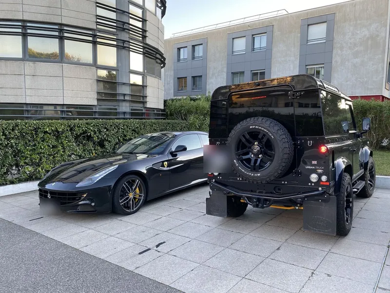 Une Ferrari et le Defender vu de dos, garés côte à côte, plaques floutées.