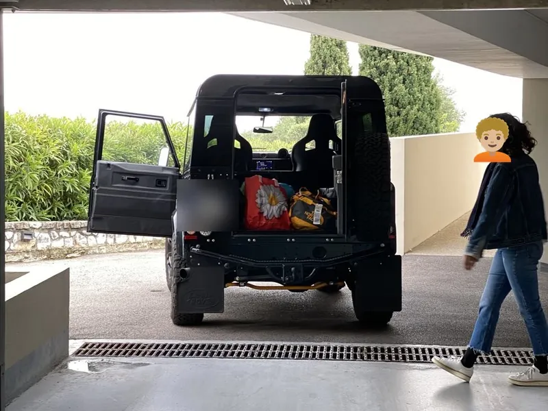 Le Defender coffre ouvert à la sortie d'un parking, plaque floutée ; une personne passe à côté, le visage remplacé par un emoji.
